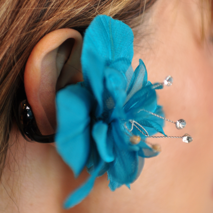 Close-up of a teal PRIAMBLE Sonnet orchid audio earring, showing layered petals, crystal accents, and the black ear clip.