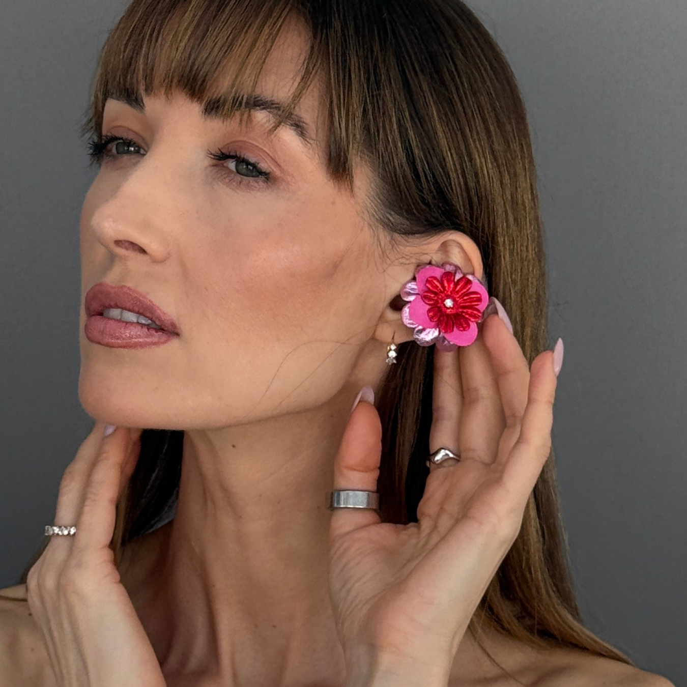 A close-up of a woman wearing a vibrant pink Priamble Audio Earrings on her ear, with her hands gently touching her face and neck against a solid gray background.