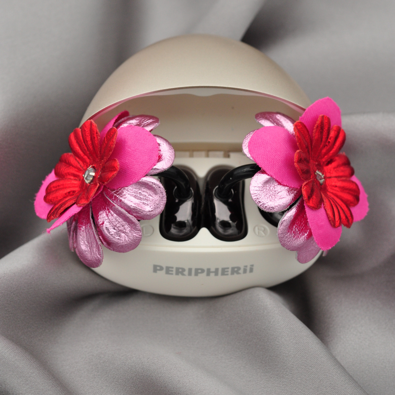 A close-up of a PERIPHERii Priamble wearable device with two large decorative fabric flowers in pink, red, and metallic pink attached to the sides, set against a draped gray fabric background.